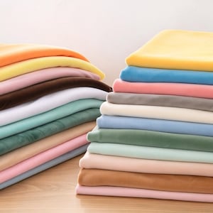 Puede incluir: Dos pilas de tela doblada en varios colores, incluyendo amarillo, naranja, rosa, marrón, blanco, azul claro y verde. La tela tiene una textura suave y lisa, dispuesta en pilas ordenadas y superpuestas sobre una superficie de madera clara.