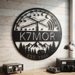 May include: A circular black metal wall sign featuring a cutout design of a radio antenna, stars, a crescent moon, a mountain range, and a small cabin. The text K7MOR is centred horizontally, with the text EST. 1986 positioned at the bottom.