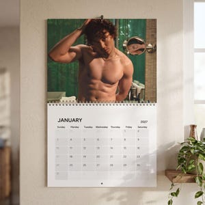 May include: A 2027 wall calendar for the month of January featuring a photograph of a shirtless person with curly hair touching their head, positioned above a white grid calendar page with black text and a spiral binding at the top.