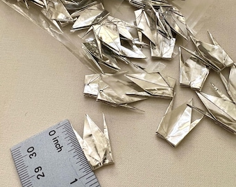 May include: A collection of small, metallic silver origami cranes made from foil paper, partially contained in a clear plastic bag. A metal ruler is positioned next to one crane, showing it measures approximately 1 inch in height.