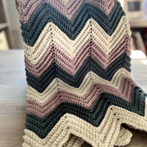 Può includere: Una coperta all'uncinetto fatta a mano con un motivo a zig-zag. La coperta presenta file alternate di crema, rosa antico, verde acqua scuro e grigio. La coperta è piegata su un cestino, mostrando la consistenza e le variazioni di colore.
