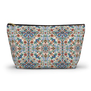 Peut inclure: Une pochette zippée présentant un motif répétitif de type carrelage avec des motifs floraux et géométriques complexes dans des tons de rouge, bleu, vert et jaune sur fond blanc. Le sac possède une tirette de fermeture éclair dorée.