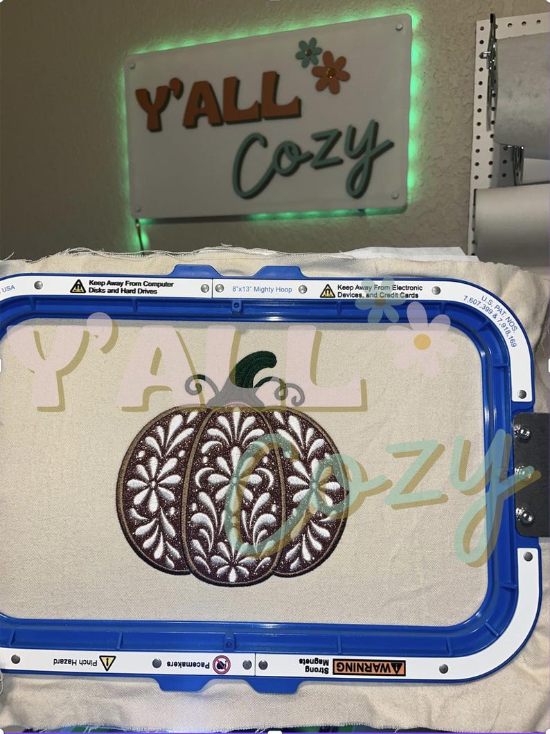 以下が含まれることがあります： 20.32 x 33.02 cmの青い刺繍枠に、ダークブラウンのグリッター仕上げと白い花柄が刺繍されたカボチャが施されたオフホワイトの布が張られています。枠の上にはY'ALL COZYという文字が書かれた看板があります。