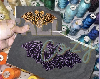 以下が含まれることがあります： 刺繍されたコウモリのデザインが施された2枚の長方形のグレーの布製パッチ。上のパッチには花のような翼の模様を持つオレンジ色のコウモリが、下のパッチには同様の細かい翼の刺繍が施された紫色のコウモリが描かれています。