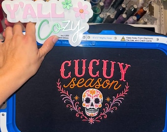 以下が含まれることがあります： 青い刺繍枠に固定された黒い布地。ピンクとオレンジで刺繍された「CUCUY season」の文字と、花柄のアクセントが付いたカラフルなシュガースカルのグラフィックが特徴。上部の白い看板には「Y'ALL Cozy」と花のアイコンが描かれている。