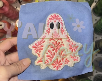 以下が含まれることがあります： 中央に刺繍された幽霊のアップリケが付いた正方形のライトブルーの布地。幽霊は白で、複雑なピンクの花の刺繍が施されており、2つの黒い楕円形の目があります。右側には小さなライトブルーの花が刺繍されています。