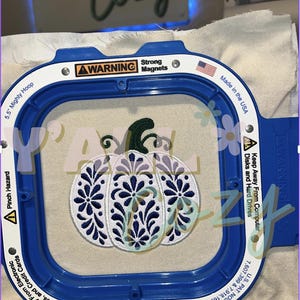 以下が含まれることがあります： 14cmの青いMighty Hoop刺繍枠に、花柄と濃い緑色の茎が描かれた濃紺と白のパンプキンデザインの白い布が固定されています。枠には磁石や電子機器に関する警告文が記載されています。