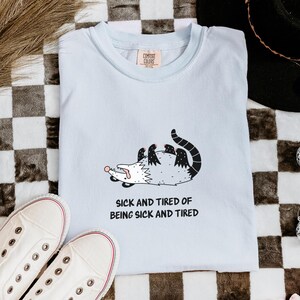 Könnte beinhalten: Ein hellblaues T-Shirt mit einer Grafik eines weiß-schwarzen Opossums, das auf dem Rücken liegt, und dem darunter in Schwarz gedruckten Text "SICK AND TIRED OF BEING SICK AND TIRED". Das Shirt hat einen Rundhalsausschnitt und ein Markenetikett.