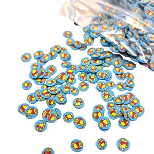 May include: A collection of small, round, light blue polymer clay slices featuring a yellow bear character wearing a red shirt. The slices are scattered across a surface, with a portion remaining inside a clear plastic resealable bag.