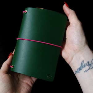 Może przedstawiać: Ciemnozielony skórzany notatnik z różowym elastycznym zapięciem. Notatnik jest trzymany na czarnym tle. Inicjały "JFR" są wytłoczone w lewym dolnym rogu notatnika.