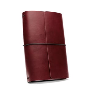 Può includere: Un diario in pelle bordeaux con chiusura a fascia elastica nera. Il diario è rettangolare e sembra chiuso. La copertina è liscia e i bordi sono arrotondati. Lo sfondo è bianco.