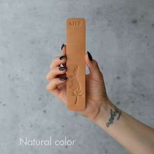 May include: A brown leather bookmark with a floral design and the initials "MEF" embossed on the top. The bookmark is held in a hand with black nail polish.