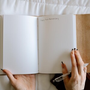 Pode incluir: Um di&aacute;rio branco em branco com uma capa de couro marrom. O di&aacute;rio est&aacute; aberto em uma p&aacute;gina em branco com as palavras "Happy First Anniversary" escritas com tinta preta.