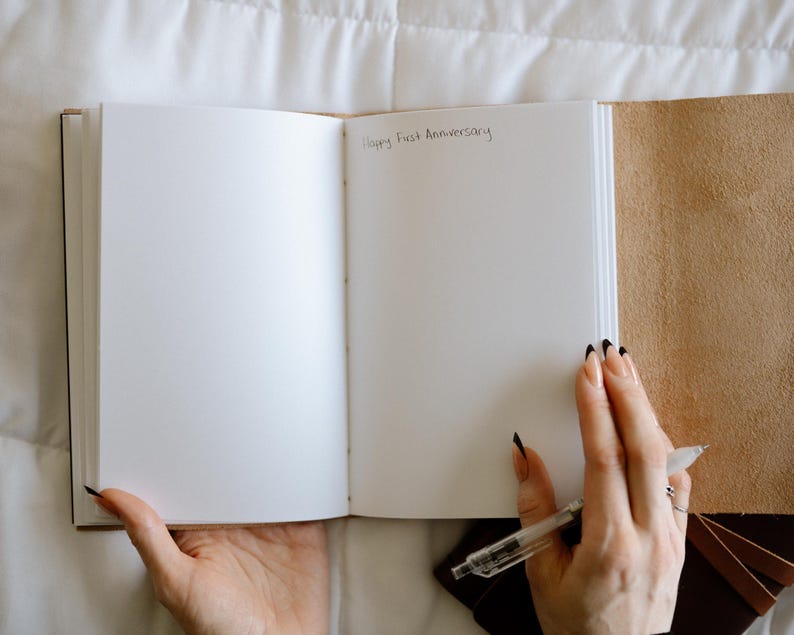 Pode incluir: Um di&aacute;rio branco em branco com uma capa de couro marrom. O di&aacute;rio est&aacute; aberto em uma p&aacute;gina em branco com as palavras "Happy First Anniversary" escritas com tinta preta.