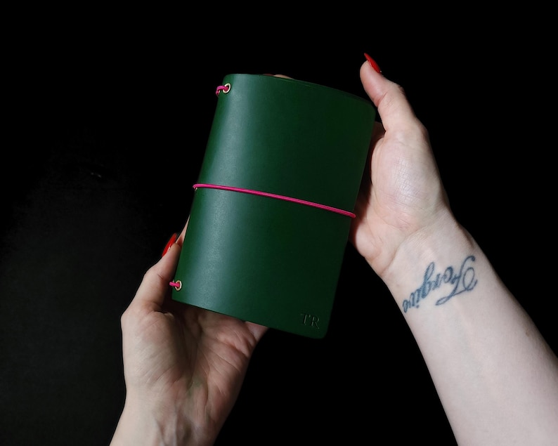 May include: A dark green leather journal with a pink elastic closure. The journal is held against a black background. The initials "JFR" are embossed on the lower left corner. The journal is approximately 5 inches tall.