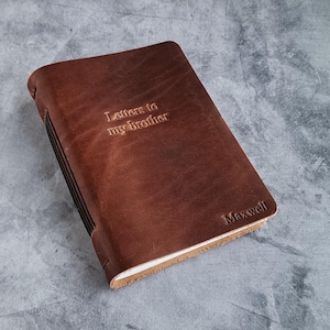 Peut inclure: Un journal en cuir marron avec le texte en relief "Letters to my brother" et le nom "Maxwell". Le journal est fermé, avec des coutures visibles le long du dos et un bord de couleur claire.