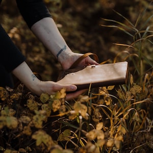 May include: A brown leather journal with a strap closure, held in the hands of a person. The journal is in a field of tall grass and leaves.