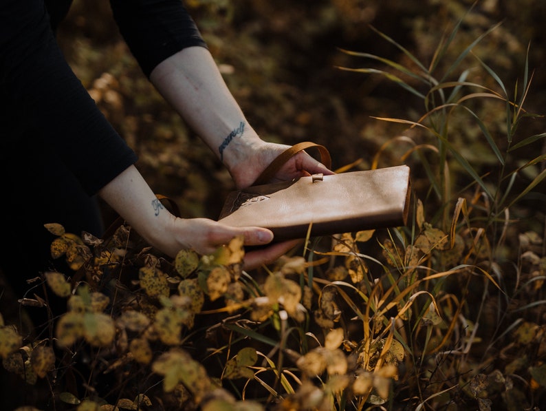 May include: A brown leather journal with a strap closure, held in the hands of a person. The journal is in a field of tall grass and leaves.