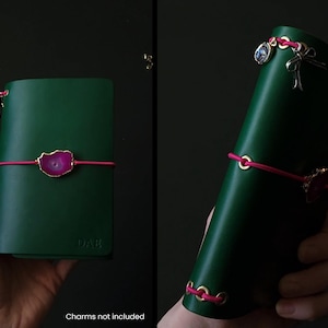 May include: A dark green leather journal with a pink elastic closure and a decorative pink and gold stone accent. The journal is held in a hand and has gold-colored eyelets. The text "DAE" is embossed on the cover.
