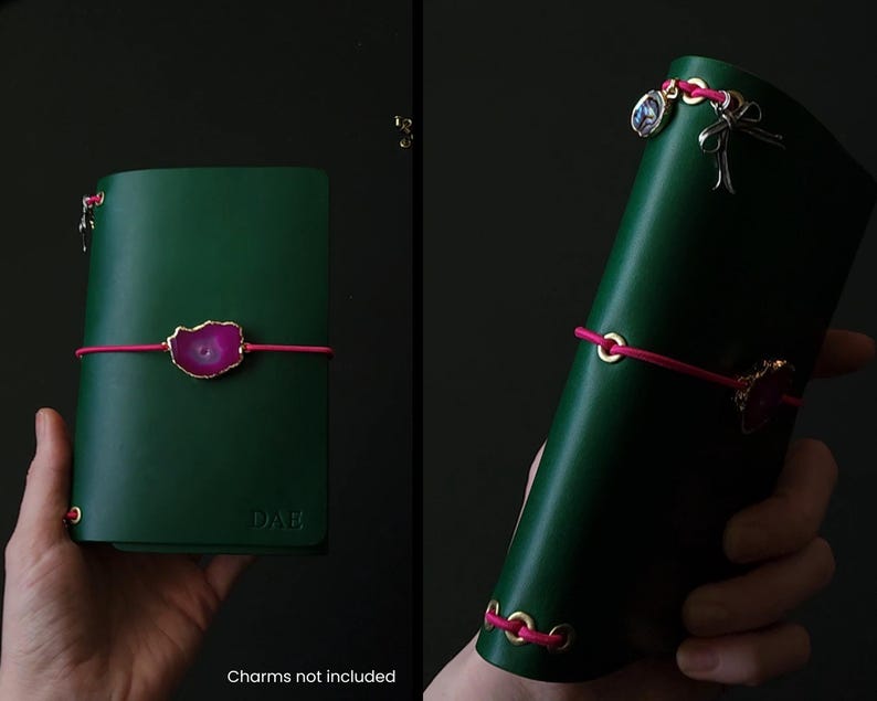 May include: A dark green leather journal with a pink elastic closure and a decorative pink and gold stone accent. The journal is held in a hand and has gold-colored eyelets. The text "DAE" is embossed on the cover.