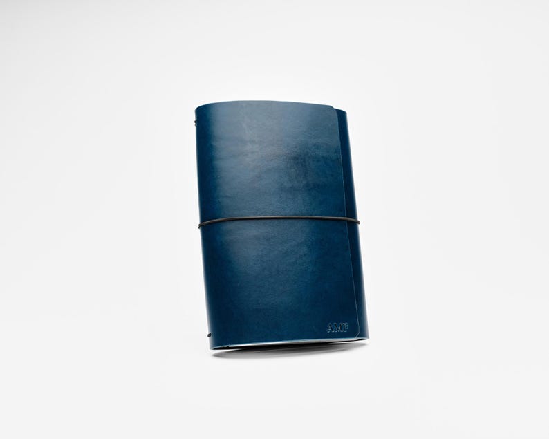 May include: A navy blue leather journal with a black elastic closure. The journal is closed and has a minimalist design. The bottom right corner has the embossed text "AM".