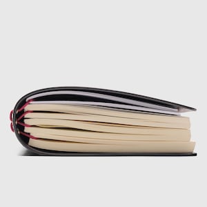 May include: A black notebook with a flexible cover and multiple stacks of cream-colored paper pages. The spine is bound with bright magenta elastic cord threaded through gold-toned metal eyelets, securing the paper sections together.
