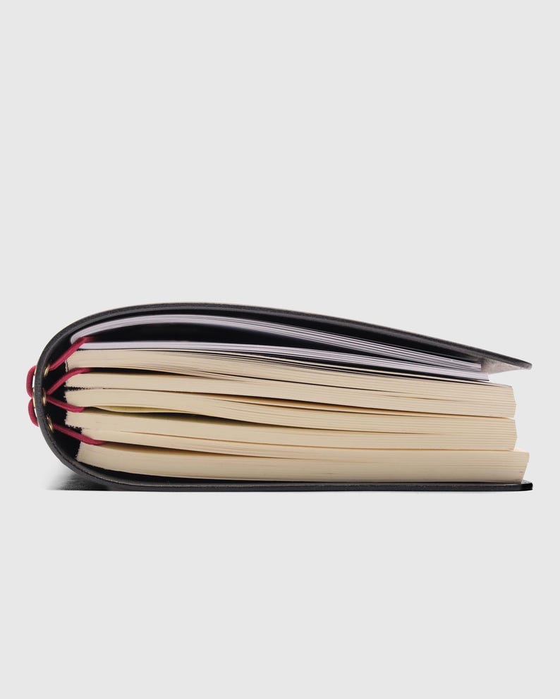May include: A black notebook with a flexible cover and multiple stacks of cream-colored paper pages. The spine is bound with bright magenta elastic cord threaded through gold-toned metal eyelets, securing the paper sections together.