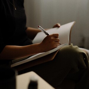 May include: A person writing in a notebook with a pen. The notebook is open and the person is holding the pen in their right hand. The person is wearing a black shirt and has their legs crossed.