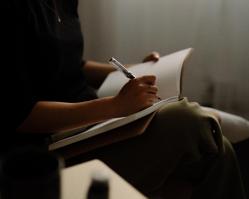 May include: A person writing in a notebook with a pen. The notebook is open and the person is holding the pen in their right hand. The person is wearing a black shirt and has their legs crossed.