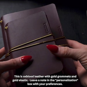 May include: Oxblood leather journal with gold grommets and elastic. The word "emma" is embossed on the cover. The journal is held together with a gold elastic band.