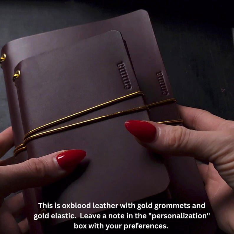 May include: Oxblood leather journal with gold grommets and elastic. The word "emma" is embossed on the cover. The journal is held together with a gold elastic band.
