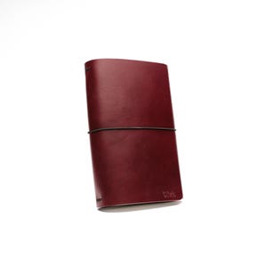 May include: A burgundy leather journal with a black elastic closure. The journal is rectangular and features a debossed logo in the lower right corner. The cover is smooth and the edges are rounded.