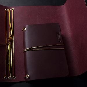May include: A dark brown leather journal with the name "emma" embossed on the cover. The journal is secured with a gold string and is set against a deep red leather background. The journal is open, revealing the interior pages.