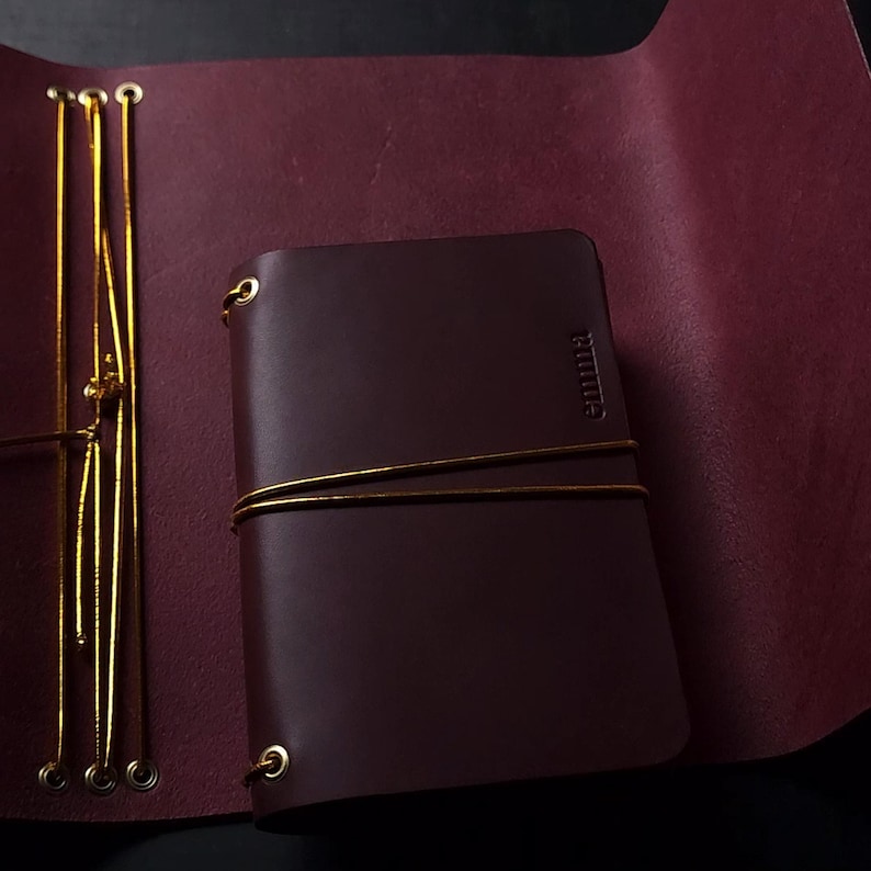 May include: A dark brown leather journal with the name "emma" embossed on the cover. The journal is secured with a gold string and is set against a deep red leather background. The journal is open, revealing the interior pages.