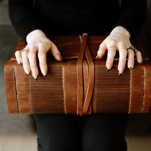 Peut inclure: Un journal relié en cuir marron avec une fermeture à boucle décorative. Le journal est fermé et repose sur les genoux d'une personne.