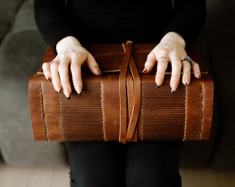 May include: A brown leather-bound journal with a decorative strap closure. The journal is closed and resting on a person's lap.