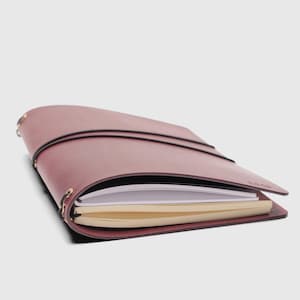 May include: A maroon leather journal cover featuring a wrap-around black elastic closure and metal eyelets along the spine. The cover holds two separate notebooks, one with white pages and one with cream-colored pages, secured by internal elastic bands.