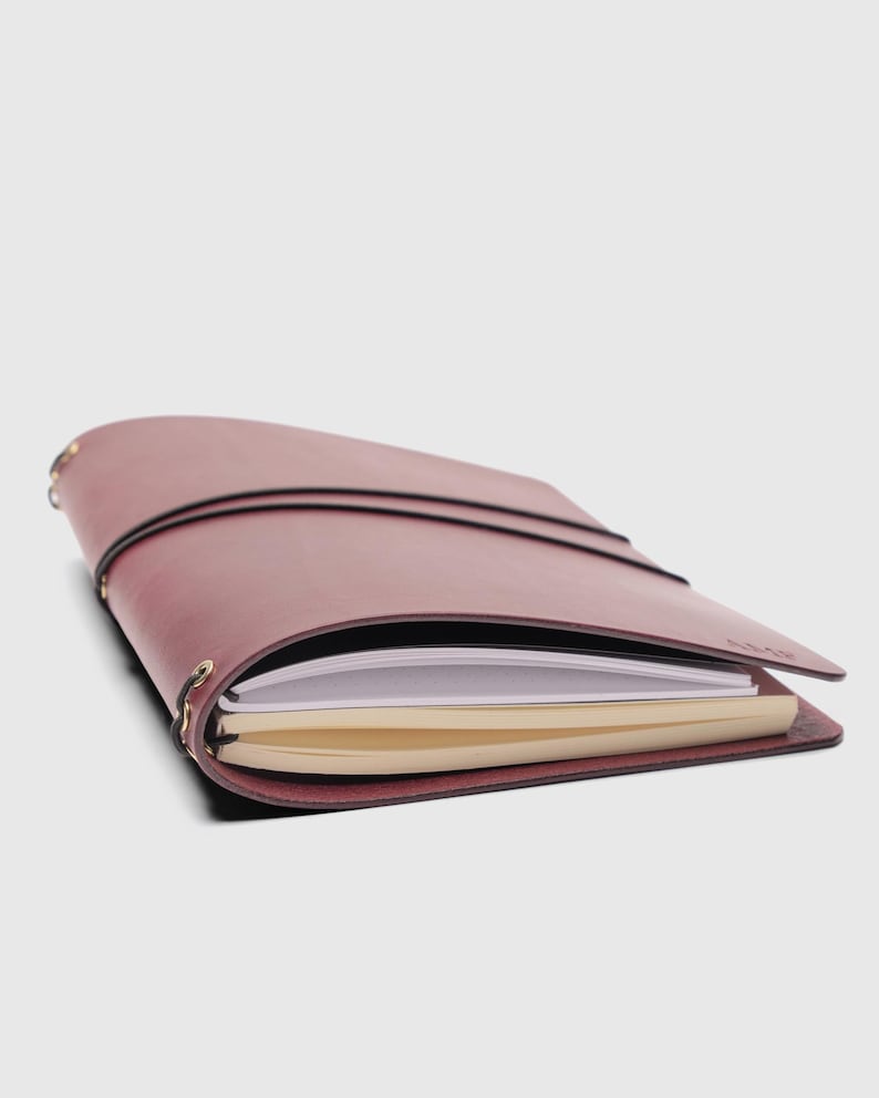 May include: A maroon leather journal cover featuring a wrap-around black elastic closure and metal eyelets along the spine. The cover holds two separate notebooks, one with white pages and one with cream-colored pages, secured by internal elastic bands.