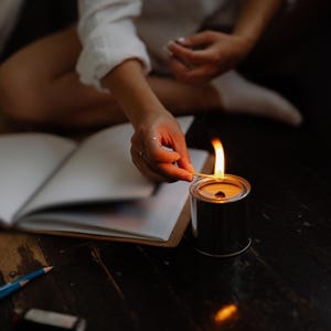 May include: A person lights a candle with a match. The candle is in a silver tin container. The person is sitting at a table with a notebook open in front of them.