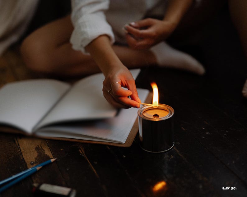 May include: A person lights a candle with a match. The candle is in a silver tin container. The person is sitting at a table with a notebook open in front of them.