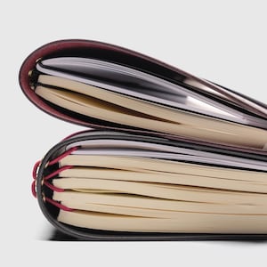 May include: Two stacked notebooks with dark covers and cream-colored pages. The top notebook has a deep burgundy spine, while the bottom notebook features a black cover secured with bright pink elastic cords threaded through gold-toned eyelets.