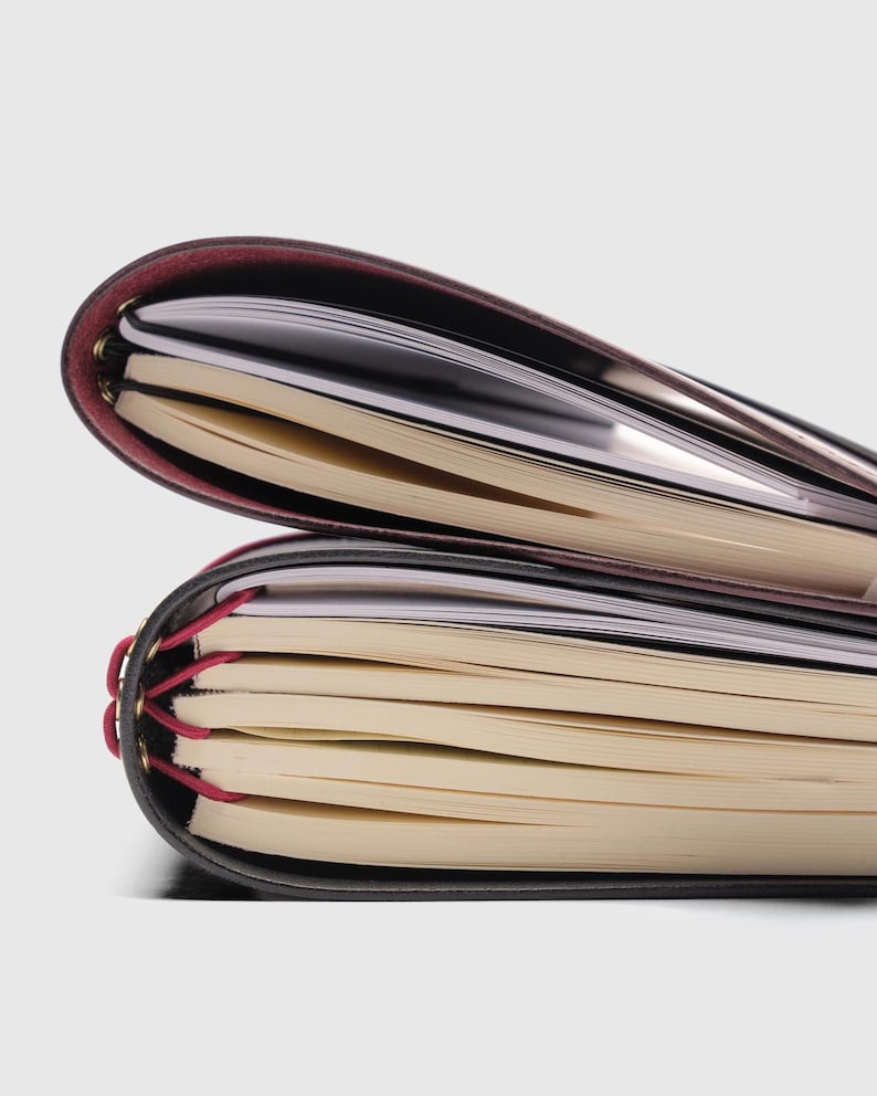 May include: Two stacked notebooks with dark covers and cream-colored pages. The top notebook has a deep burgundy spine, while the bottom notebook features a black cover secured with bright pink elastic cords threaded through gold-toned eyelets.