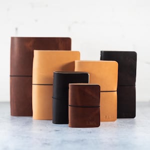 May include: A collection of six leather-bound journals in varying sizes and shades of brown, tan, and black. Each notebook is secured with a thin black elastic band wrapped horizontally around the cover. Some journals feature embossed initials.