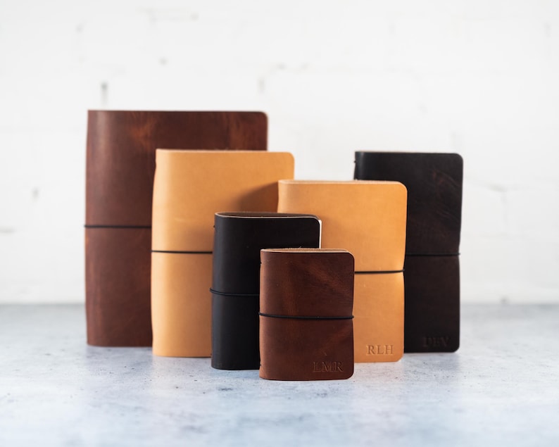 May include: A collection of six leather-bound journals in varying sizes and shades of brown, tan, and black. Each notebook is secured with a thin black elastic band wrapped horizontally around the cover. Some journals feature embossed initials.