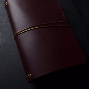 May include: A burgundy leather journal with gold-colored metal accents. The journal is secured with a gold-colored elastic band. The word "Ruitert" is embossed on the cover. The journal is closed and appears to be new.