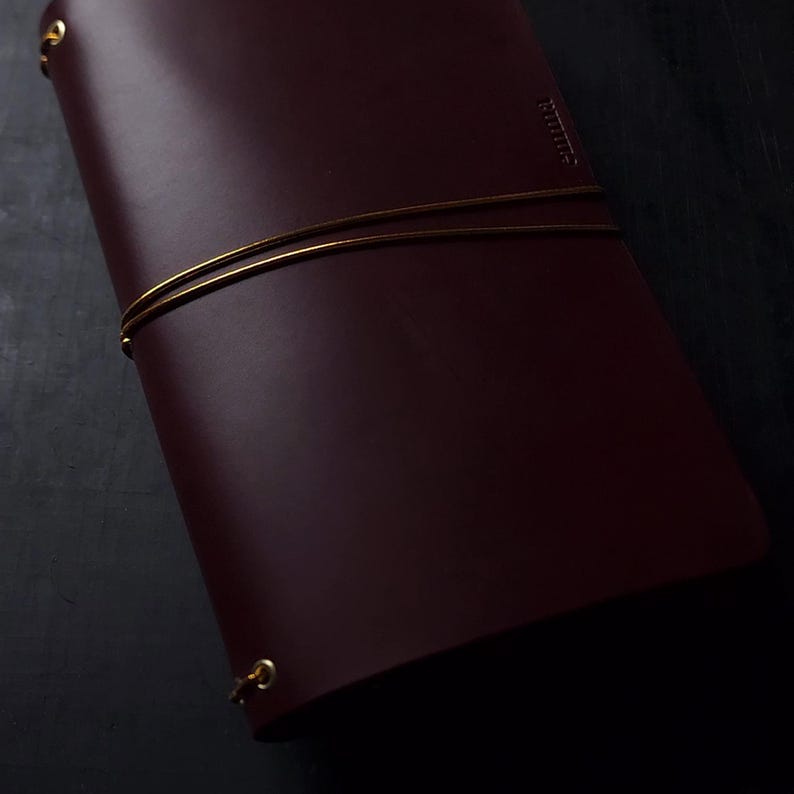 May include: A burgundy leather journal with gold-colored metal accents. The journal is secured with a gold-colored elastic band. The word "Ruitert" is embossed on the cover. The journal is closed and appears to be new.