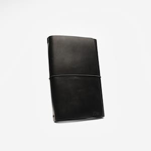 May include: A black leather journal with a simple design. The journal is rectangular, with a black elastic band closure. The cover is smooth and the edges are rounded. The journal is on a white background.