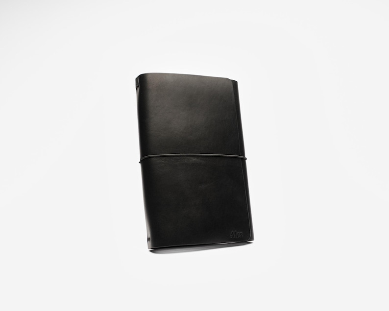 May include: A black leather journal with a simple design. The journal is rectangular, with a black elastic band closure. The cover is smooth and the edges are rounded. The journal is on a white background.