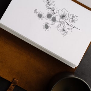 May include: A brown leather journal with a white page open to reveal a black and white line drawing of a bouquet of flowers.