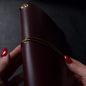 May include: A burgundy leather journal with a gold-colored elastic closure. The journal is closed, with the gold elastic wrapped around it. The journal's cover is smooth and has a slight sheen. The background is dark.